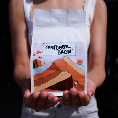 Person holding a coffee bag labeled 'Outdoor Brew' with a sand dune scenic design. Coffee is the roasters blend, caramel dunes.