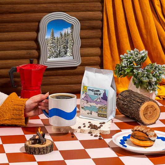 Person holding a coffee mug with a scenic backdrop, featuring a coffee bag and snacks on a checkered table. scene is set inside a lodge with a warm and cozy vibe matching the coco tones of the coffee 