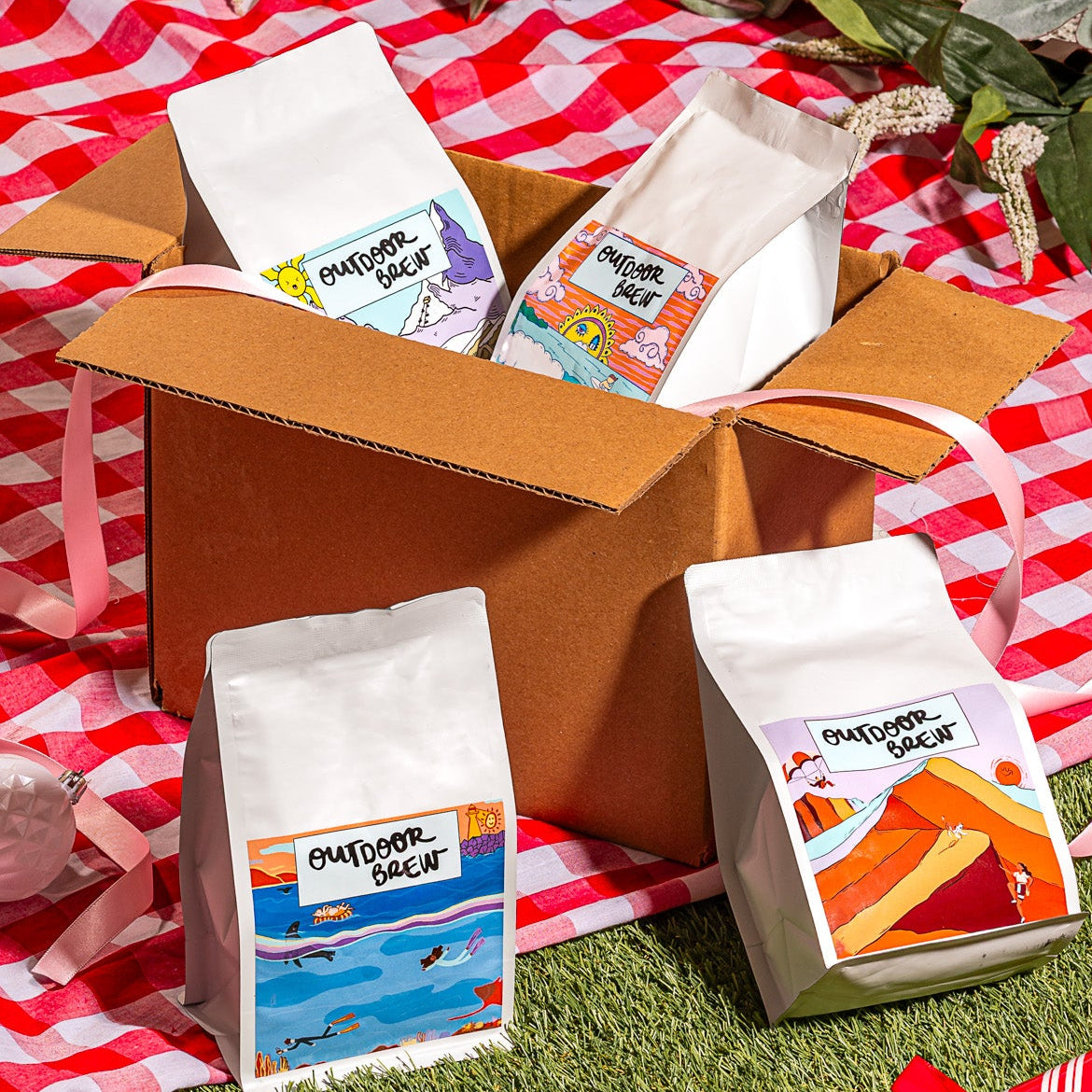 Box of outdoor 250g coffee packets on a red and white checkered picnic blanket with decorative elements.