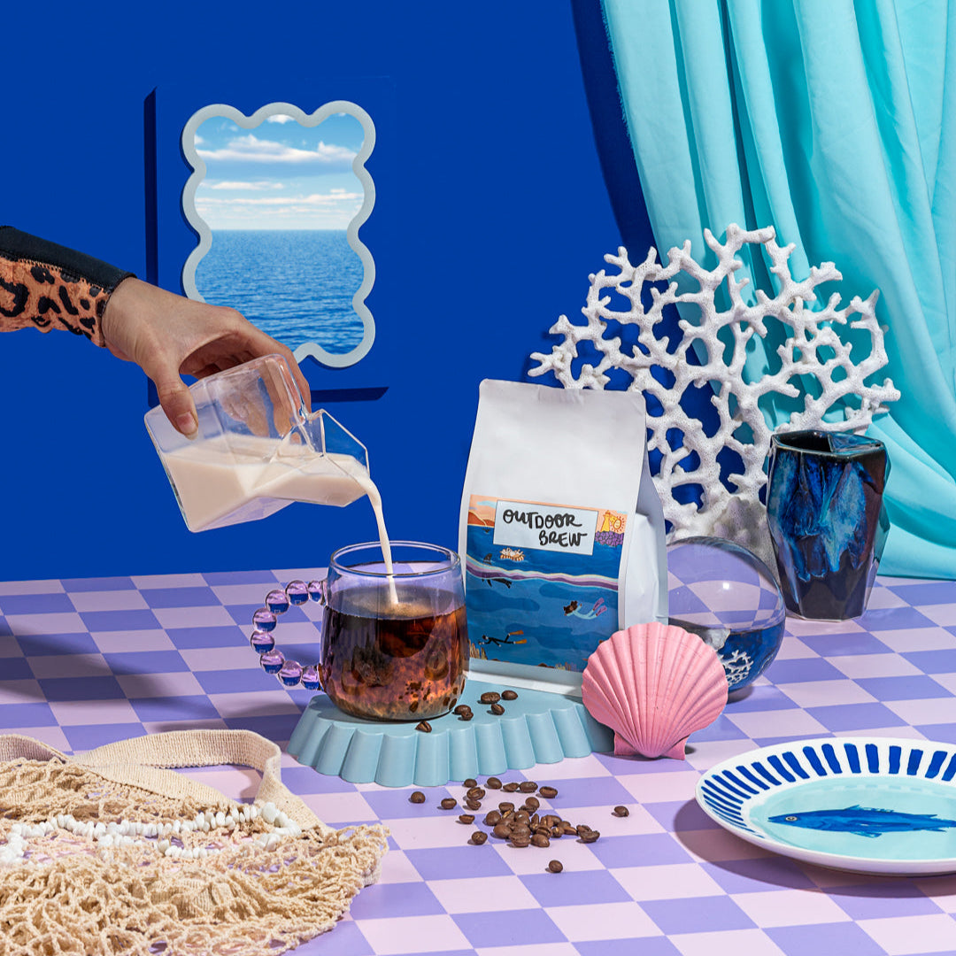 Person pouring milk into a glass of coffee on a table with decorative items and a blue curtain. Dark harbour Sumatran coffee beans