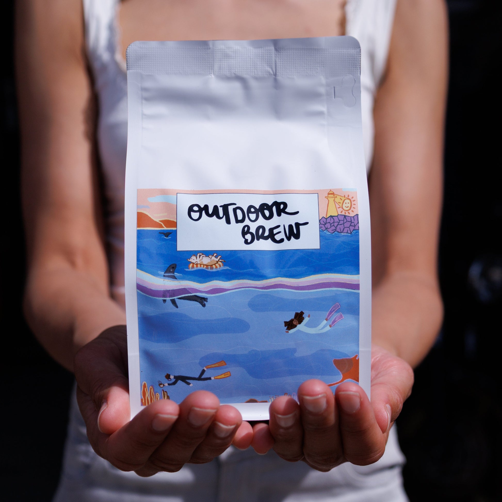 Person holding a package labeled 'Outdoor Brew' with an under water design. the coffee is dark harbour, their brazil single origin