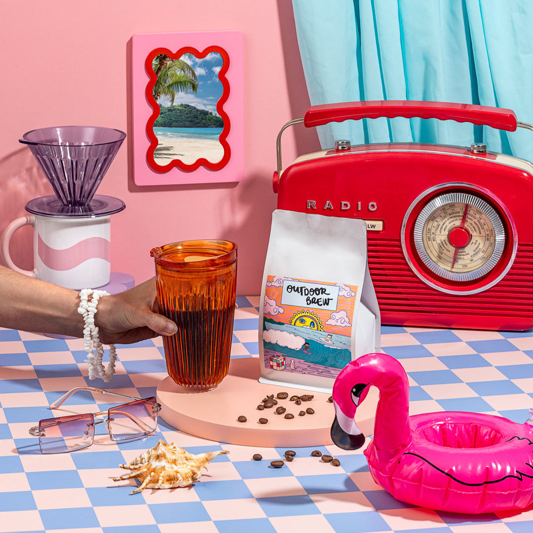 Vintage-style radio, coffee cup, and flamingo floatie on a checkered tablecloth with a pink wall and framed picture. this is sunset shores, outdoor brew's single origin PNG coffee