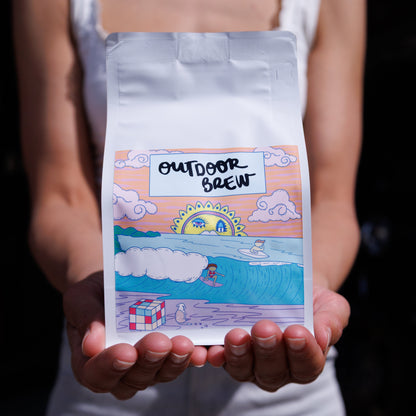 Person holding a bag of 'Outdoor Brew' coffee with a scenic ocean label. It is a cool cartoon style drawing of maroubra beach. the coffee is Sunrise Shores,  a PNG single origin